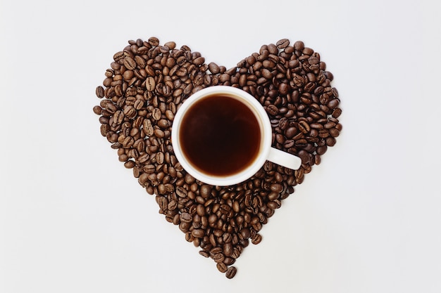 Granos de café tostados en forma de corazón y taza de café en blanco