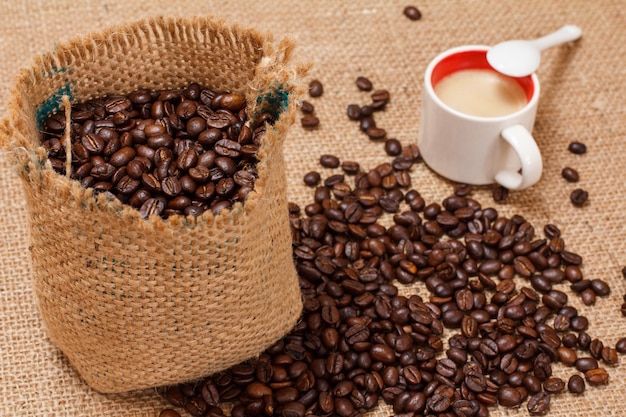 Granos de café tostados en un saco de lona sobre tela de saco con una taza de café en el fondo. Vista superior.