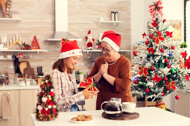 Grany mirando joygfull sobrina disfrutando de regalos navideños vistiendo sombrero rojo