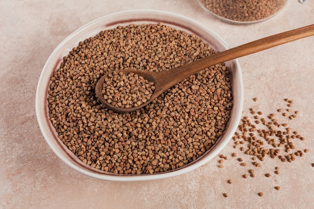Grão de trigo mourisco espalhado cru orgânico em uma tigela com uma colher de pau na superfície bege claro. Conceito de comida saudável e dieta.
