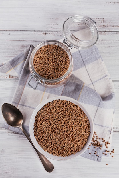 Grão de trigo mourisco espalhado cru orgânico em uma tigela e frasco de vidro no fundo rústico de madeira branco. Conceito de comida saudável e dieta. A sobrecarga