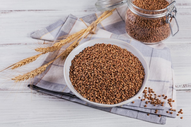 Grão de trigo mourisco espalhado cru orgânico em uma tigela e frasco de vidro sobre um fundo rústico de madeira branco. Conceito de comida saudável e dieta.