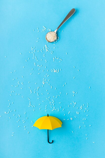 Grãos de arroz que derramam da colher no guarda-chuva amarelo do brinquedo na tabela azul. Conceito artístico de chuva de primavera