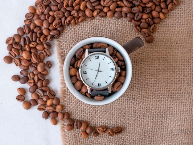 Grãos de café são espalhados em um guardanapo de lona e um relógio de mão em uma xícara de café na hora do almoço