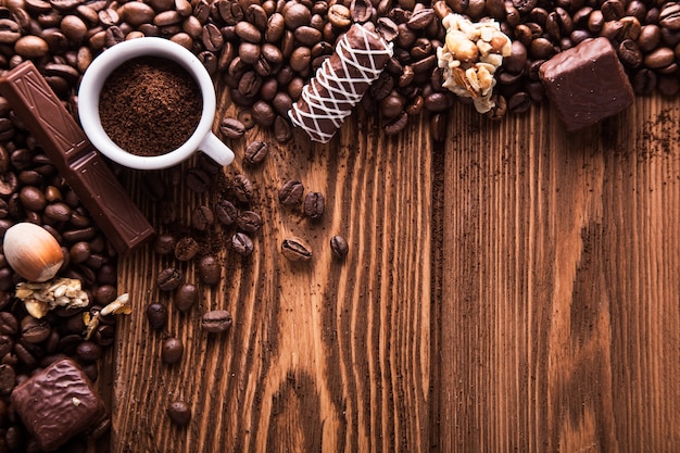 Grãos de café torrados, chocolate, doces, nozes e uma xícara com café moído na superfície de madeira