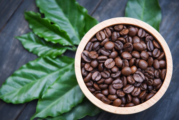 Grãos de café torrados na tigela de madeira com folha de café verde sobre fundo escuro