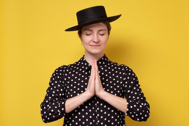 Gratefu mulher caucasiana de chapéu preto meditando orando com esperança fé juntos de mãos dadas. Estúdio filmado na parede amarela.