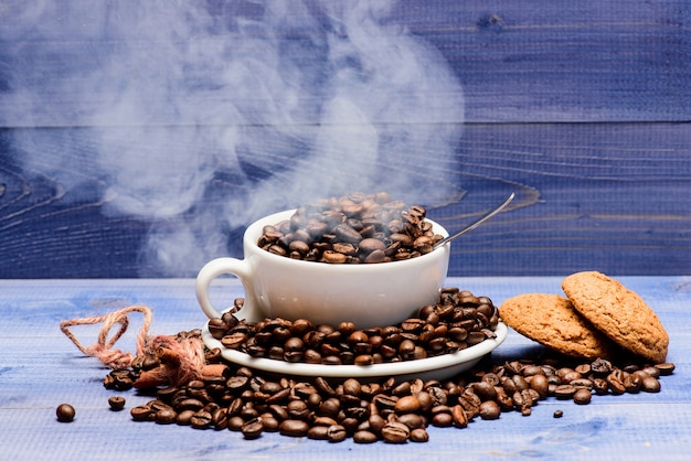 Foto grau de torra do grão. xícara de café completo marrom grãos torrados nuvens brancas de fundo de madeira de fumaça azul. menu de bebidas do café. grãos de café torrados frescos. café para inspiração e carga energética.