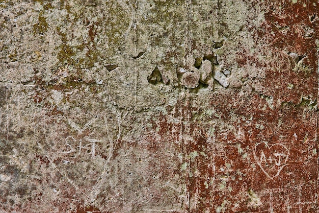 Graue Betonmauer, bedeckt mit rotem Rost und grünem Moos, mit zerfallenden Teilen und geschnitzten Herzen