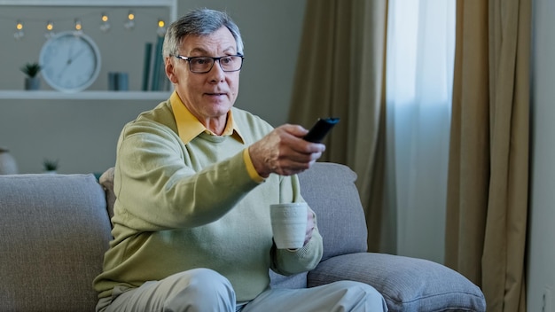 Grauhaariger alter Mann mit Brille sitzt auf der Couch vor dem Fernseher und genießt es, sich zu Hause zu entspannen und Tee zu trinken