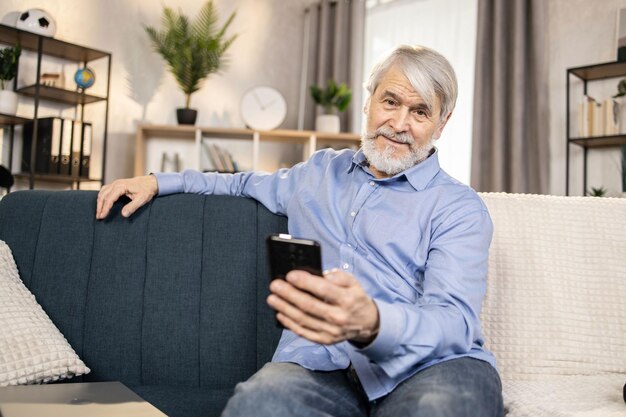 Grauhaariger Mann benutzt Telefon, während er im Wohnzimmer Platz nimmt