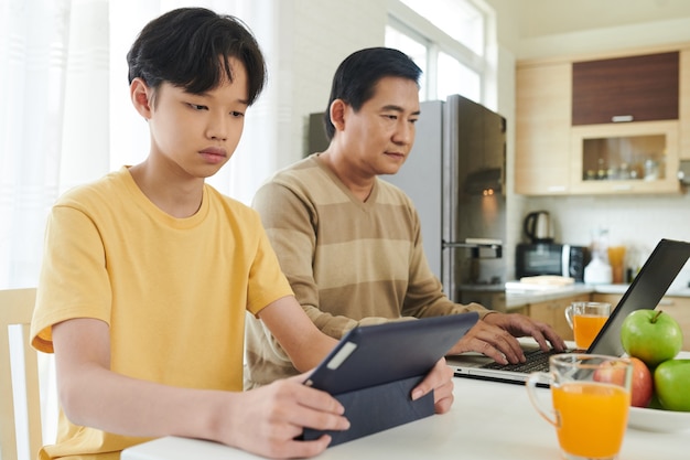 Grave adolescente leyendo libros de estudiantes en la computadora de la tableta cuando su padre trabaja en la computadora portátil junto a él