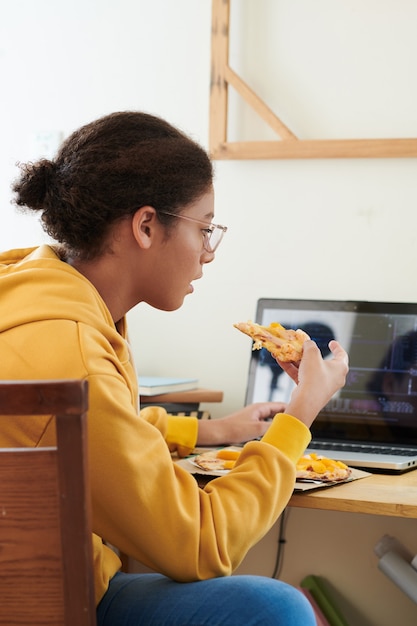 Grave adolescente de raza mixta en anteojos sentado en el escritorio y comiendo pizza mientras prepara el proyecto escolar en la computadora portátil