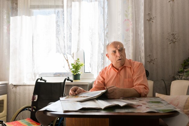 Grave anciano calvo en camisa naranja sentado en su mesa con el periódico mientras mira el marco derecho.