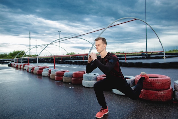 Grave atleta joven hace estocadas al aire libre. El hombre se calienta antes de correr por la mañana. El chico hace los ejercicios tirando un pie sobre el neumático. Entrenamiento al aire libre