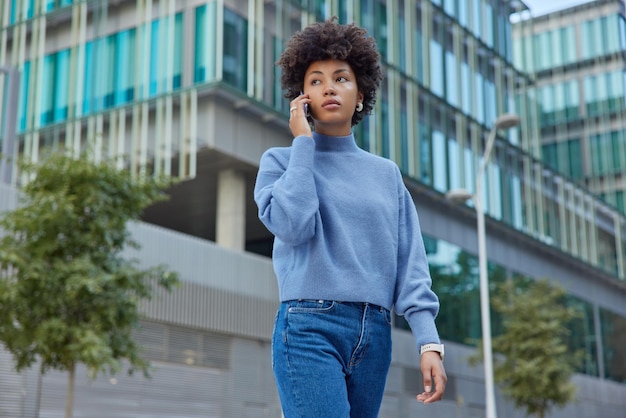 Grave bela jovem com cabelo encaracolado usa jumper casual e jeans chama alguém via smartphone tem poses de conversa contra o edifício moderno da cidade. Conceito de tecnologia de estilo de vida de pessoas