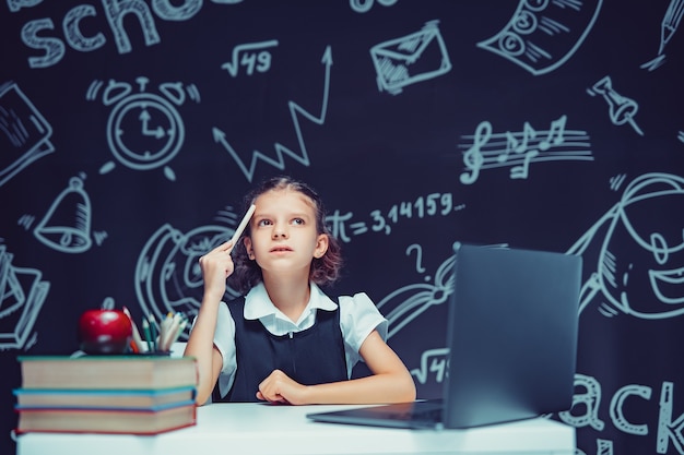 Grave colegiala reflexiva levantando la pluma sentado en el escritorio con educación a distancia portátil