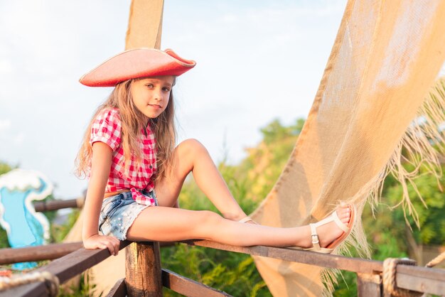 Grave divertido pirata niña con una camisa a cuadros y pantalones cortos de mezclilla se aferra a los cables del barco