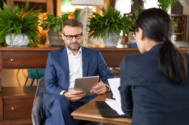 Grave empresario de mediana edad o emprendedor con touchpad mirando a través de información en línea con un colega ocupado en la parte delantera