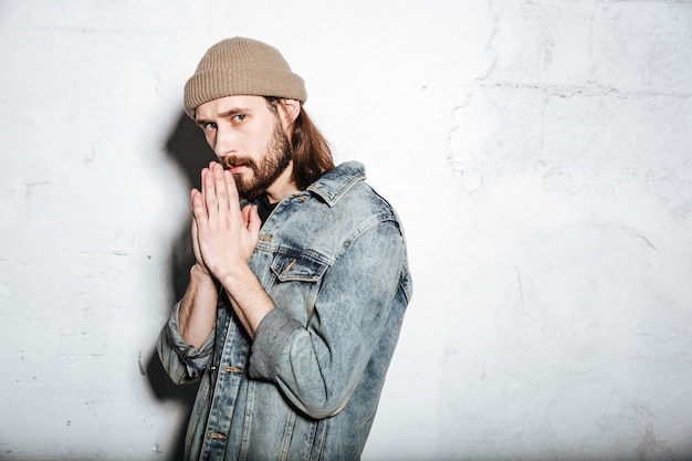 Grave hipster barbudo hombre vestido con sombrero vestido con chaqueta de jeans reza sobre la pared