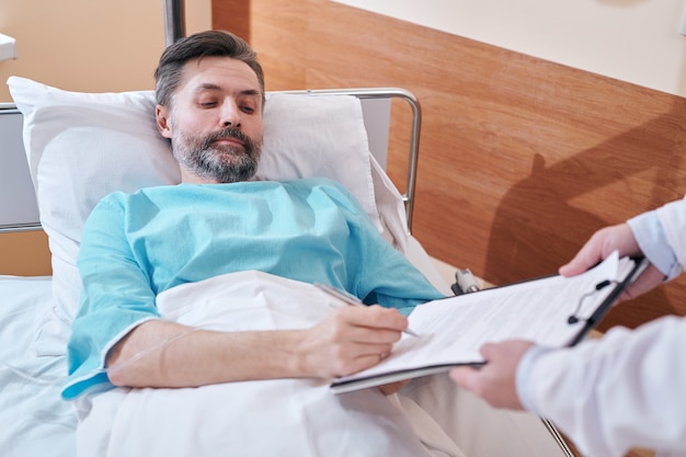 Foto grave hombre barbudo maduro en la cama de un hospital firmando un acuerdo médico antes de la operación quirúrgica