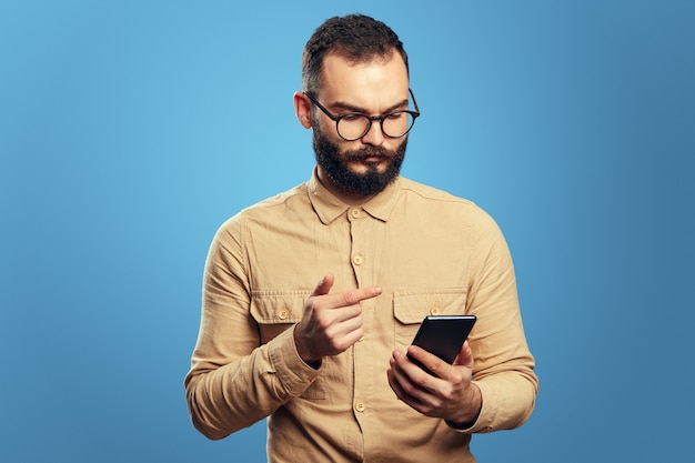 Grave hombre caucásico guapo con smartphone sobre fondo azul.