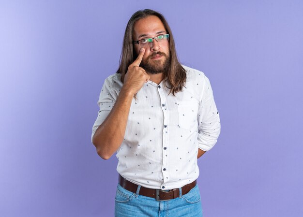 Grave hombre guapo adulto con gafas manteniendo la mano detrás de la espalda mirando a la cámara tirando hacia abajo el párpado inferior aislado en la pared púrpura con espacio de copia