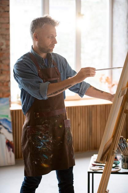 Grave hombre de mediana edad con pincel mirando la imagen en el caballete mientras pinta en su taller o estudio