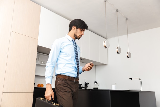 Grave hombre de negocios joven concentrado en la cocina con maleta mediante teléfono móvil.