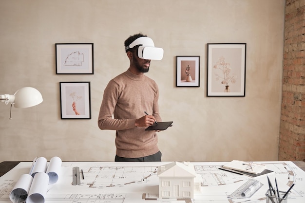 Grave jovem arquiteto afro-americano em suéter em pé no escritório moderno com fotos na parede e desenho de maquete em óculos de realidade virtual