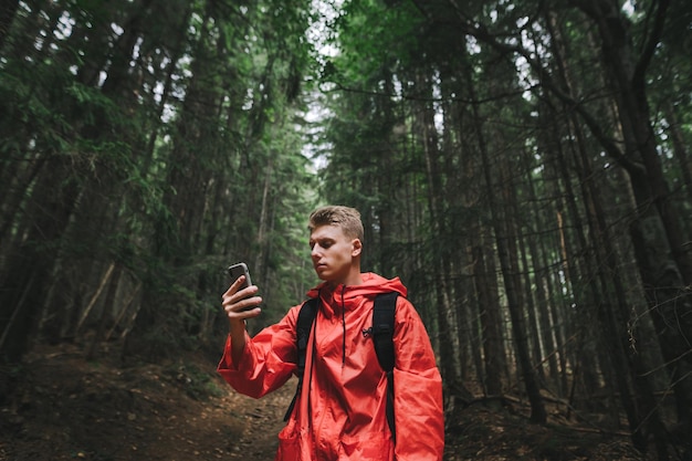 Grave jovem bonito em uma capa de chuva vermelha usa um smartphone em uma caminhada