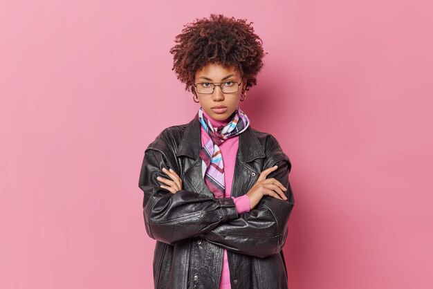 Grave jovem de cabelos encaracolados mantém os braços cruzados parece autoconfiante na câmera usa óculos jaqueta de couro e lenço amarrado no pescoço isolado sobre fundo rosa pensa em algo
