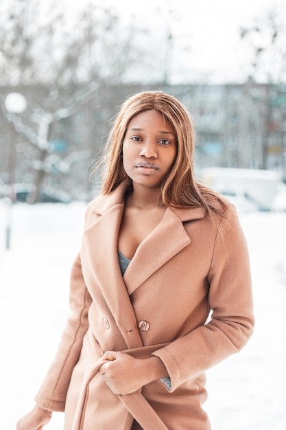 Grave jovem linda africana com um casaco bege contra o pano de fundo da neve. Mulher negra caminha na cidade