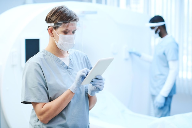 Grave jovem médica em escudo protetor facial usando tablet na sala médica com scanner de ressonância magnética