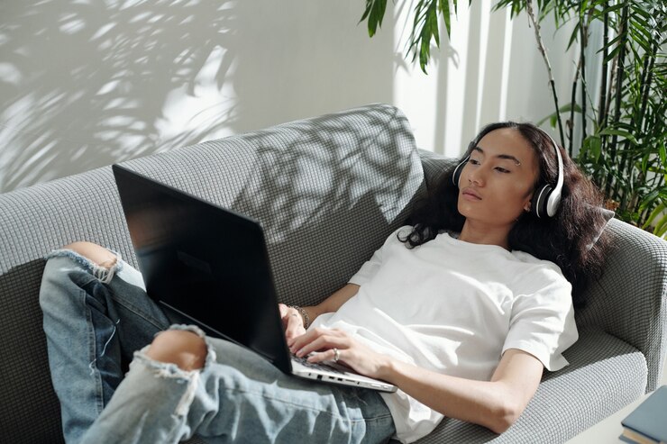 Foto grave joven asiático moderno en auriculares acostado en el sofá y usando la computadora portátil mientras trabaja con las redes sociales en casa