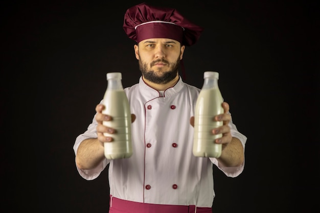 Grave joven chef barbudo en uniforme sosteniendo dos botellas de leche de plástico