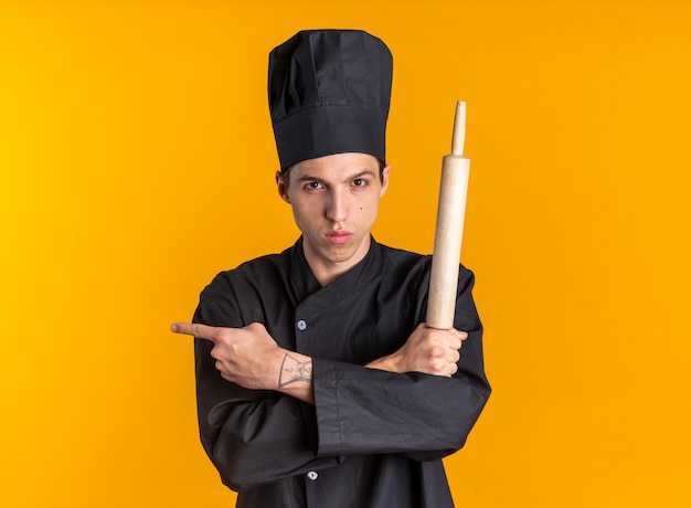 Grave joven cocinero de sexo masculino rubio en uniforme de chef y gorra manteniendo las manos cruzadas sosteniendo el rodillo apuntando al lado