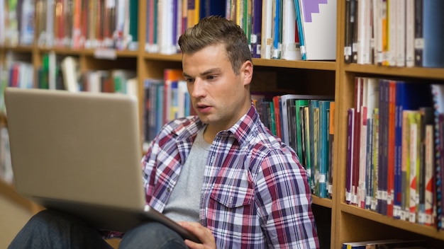 Grave joven estudiante sentado en el piso de la biblioteca usando laptop