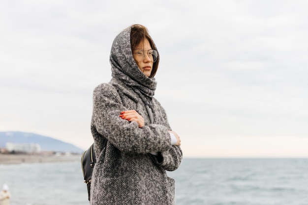 Grave joven morena con gafas y una bufanda gris se encuentra junto al mar, clima frío