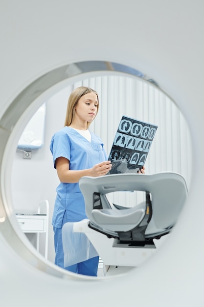 Grave joven radiólogo rubio en uniforme mirando la imagen de rayos X de la cabeza y el cerebro del paciente contra el entorno del consultorio médico en las clínicas