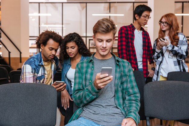 Grave joven sentado y usando el teléfono celular mientras sus amigos están de pie y hablando