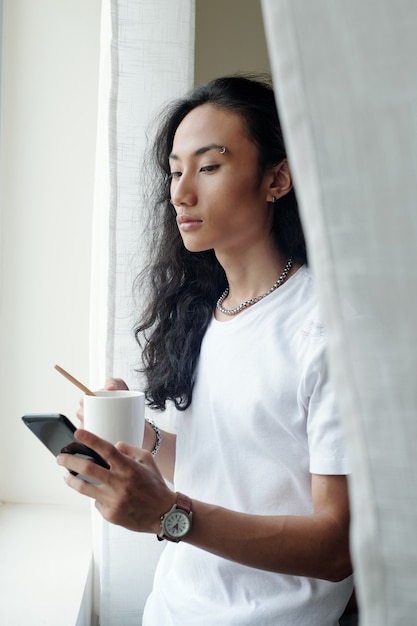 Foto grave joven vietnamita con cabello ondulado de pie en la ventana y tomando café mientras se desplaza por las redes sociales