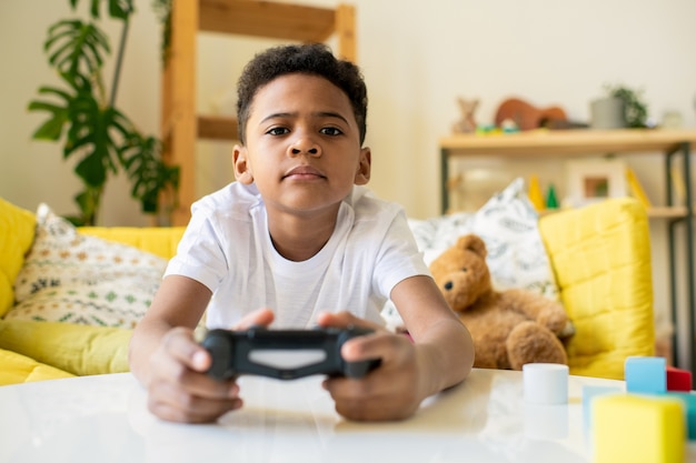 Grave lindo niño de raza mixta con joystick sentado junto a la mesa en la sala de estar