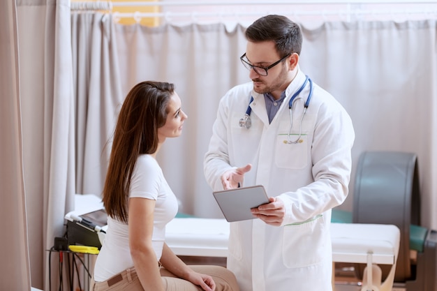 Grave médico caucásico con anteojos y uniforme azul que muestra su plan de tratamiento de paciente femenino en tableta. Interior del hospital.