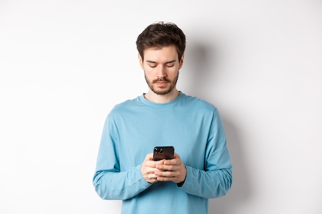 Grave mensaje de lectura joven en smartphone, mirando la pantalla del móvil, de pie sobre fondo blanco.