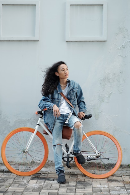 Grave modelo vietnamita joven guapo con el pelo largo en el viento sentado en bicicleta con ruedas naranjas y mirando a la distancia