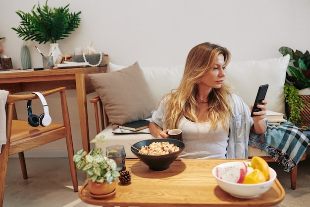 Foto grave mujer joven atractiva sentada en el suelo frente a su mesa de café, comiendo desayunos y leyendo publicaciones en las redes sociales