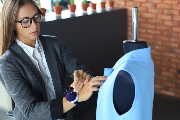 Foto grave mujer joven en gafas con agujas de coser para coser una chaqueta en maniquí mientras está de pie en su taller.