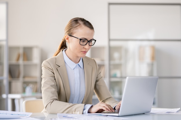 Grave mujer joven ocupada con gafas sentado en el escritorio en la oficina moderna y escribiendo en el teclado mientras prepara la presentación en la computadora portátil