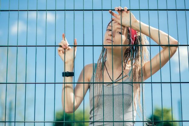 Grave mujer joven con rastas mirándote atentamente mientras está de pie en el campo de deportes cerca de la valla de tela metálica